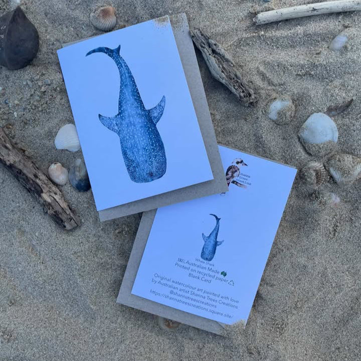 Two greeting cards with whale illustrations on a sandy surface with driftwood and shells.