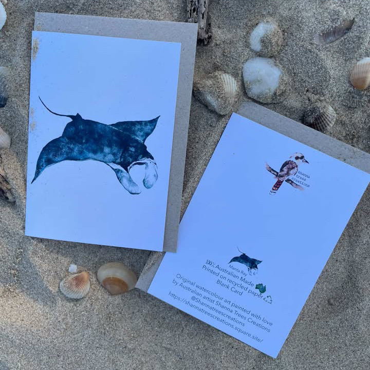 Two greeting cards with manta ray illustrations on a sandy surface with shells.