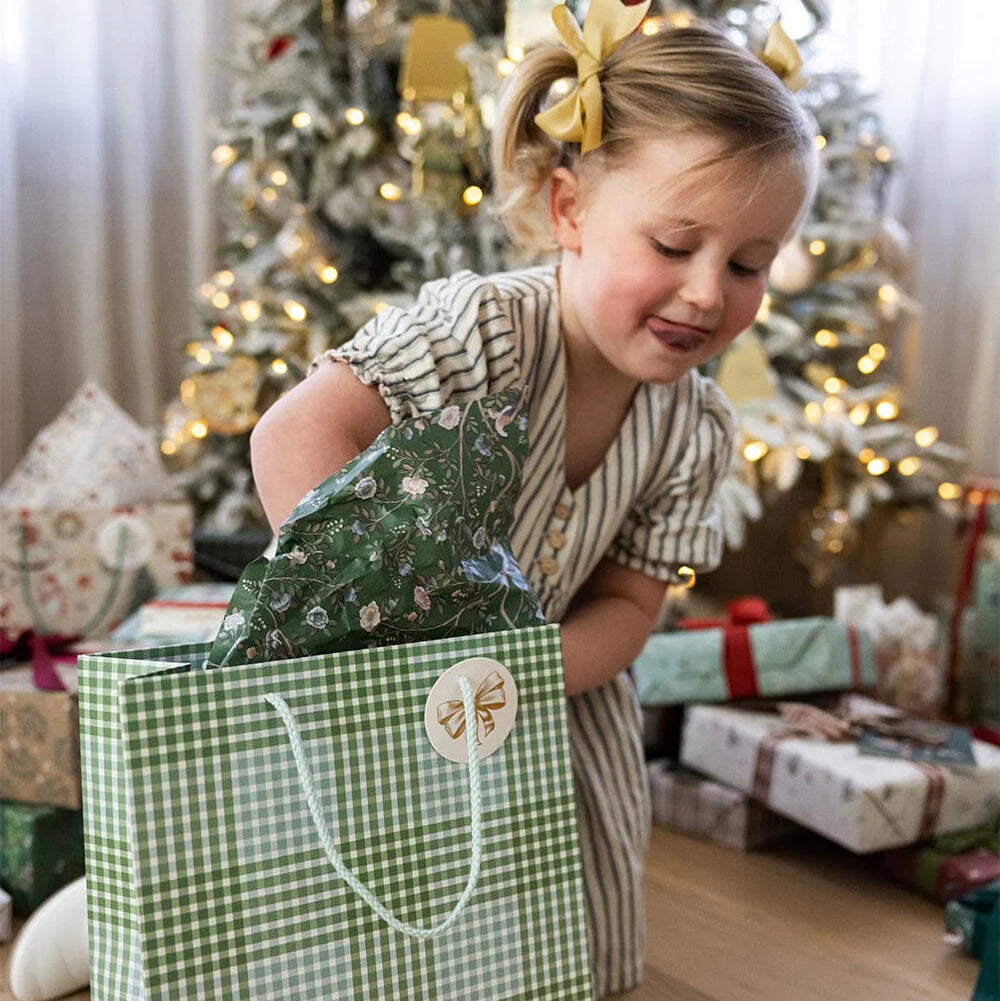 Child playing with a large green gift bag in a festive setting with Christmas trees and presents.