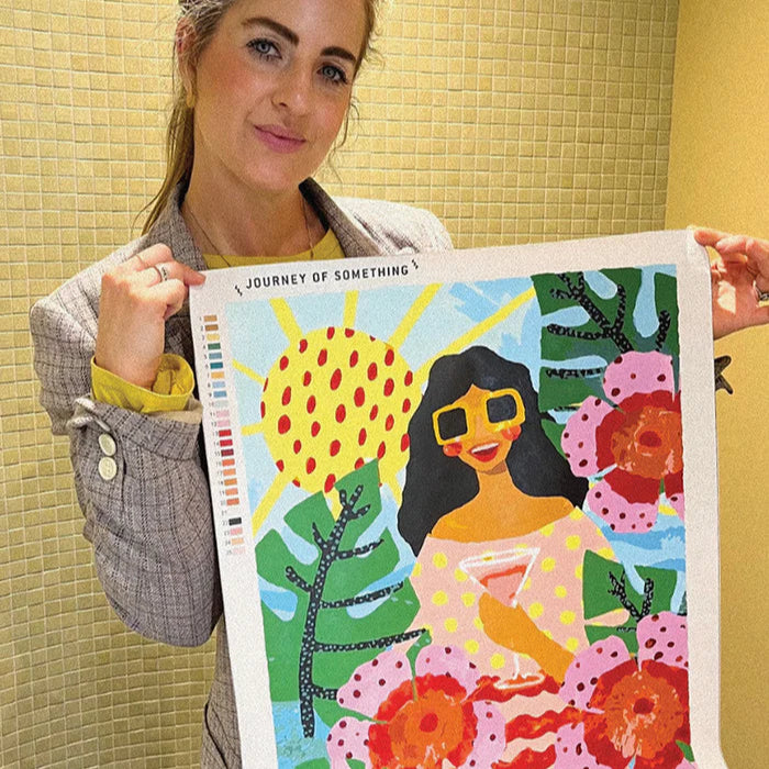 Person holding a colorful artwork with a woman, sun, and flowers on a yellow tiled wall background.